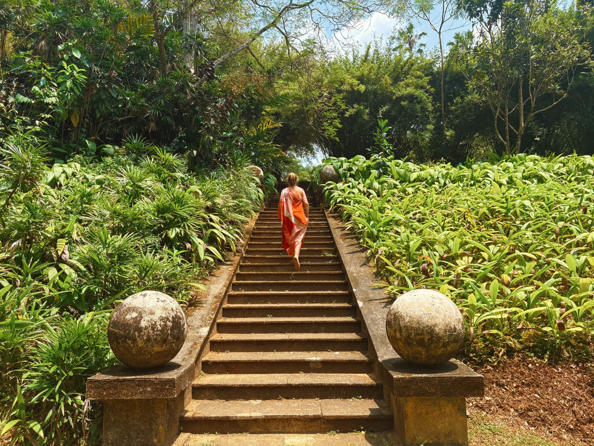 Woman in a flowing orange dress walking up stone stairs surrounded by lush green gardens